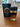 A #32 - Fahad perfume bottle by Dubai Essences, featuring a black and gold design, sits beside its matching box on a wooden table with a blurred monitor and other items in the background.