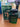 A green and black bottle of True Heritage #39 by Dubai Essences sits next to its matching box, featuring a sleek design with the brands name in white. In the blurred background, there is a computer keyboard and a smartphone on a stand.